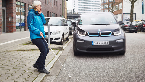 Eine Frau geht mit Langstock über die Straße, ein Auto nähert sich schnell. Im Hintergrund sind hohe Häuser und ein Baum. Die Frau hält sich erschrocken die Hand ins Gesicht.