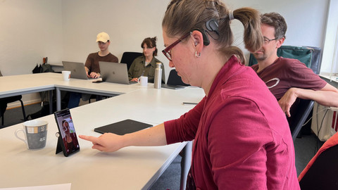 Frau Wegerhoff tippt mit dem Finger auf dem Smartphone am Tisch. 