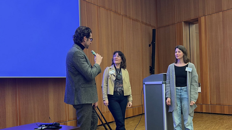 Übergabe des Testkoffers mit IKI-TAU-Materialien an das Schulministerium NRW In der Mitte steht Frau Reichstein mit dem schwarzen Testkoffer. Links von ihr steht Herr Mertens, der in das Mikrofon spricht. Rechts von ihr steht eine Frau, die die beiden anschaut.