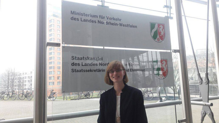 Frau Stiller steht vor dem Logo des Minsterium für Verkehr  des Landes Nordrhein Westfalen